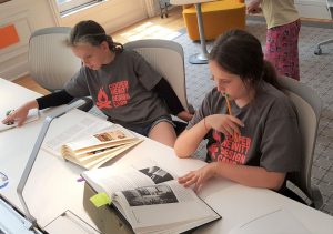 This image features 2 young girl Design campers sitting looking at library works.at table looking