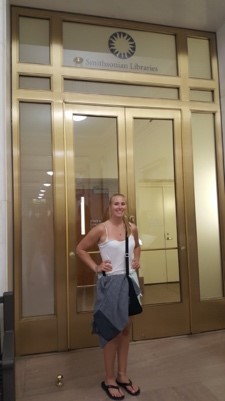 Intern Mckenna Heim standing in front of large glass and brass doors with Smithsonian Libraries signage.