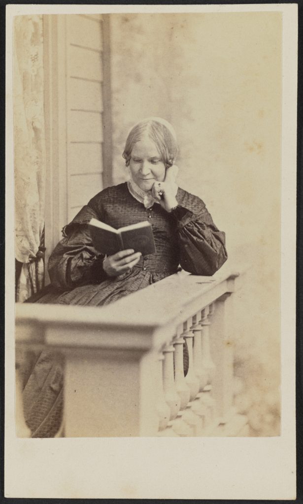 Carte-de-visite of Lydia Maria Child shown in half portrait. Child is seated behind the corner of a white columned banister, possibly on a balcony outside a house with wooden siding next to a window with a lace curtain. Her left elbow is propped on the railing and her left hand rests on her chin. She holds a book in her right hand, which she is reading. Her hair is parted at the center and gathered into curls or braided above her ears, then pulled behind her head. She wears a dark colored bodice and skirt with dropped bell sleeves and white lace collar. There is a ring with a dark stone on her left hand ring finger.