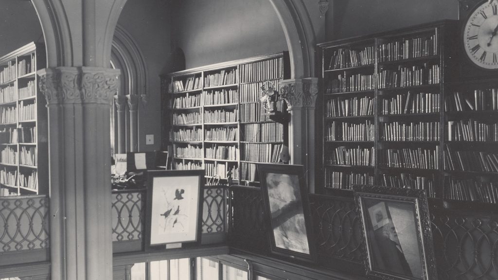 Black and white photograph of library with shelves of books and framed pictures. 