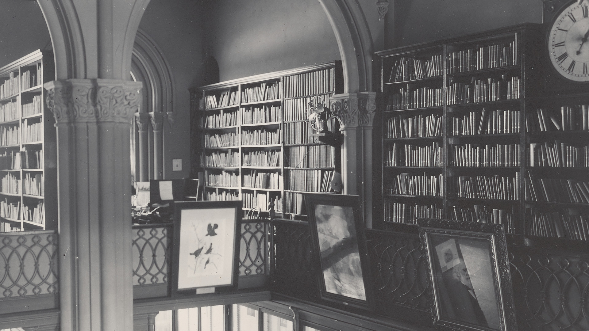 Black and white photograph of library with shelves of books and framed pictures.