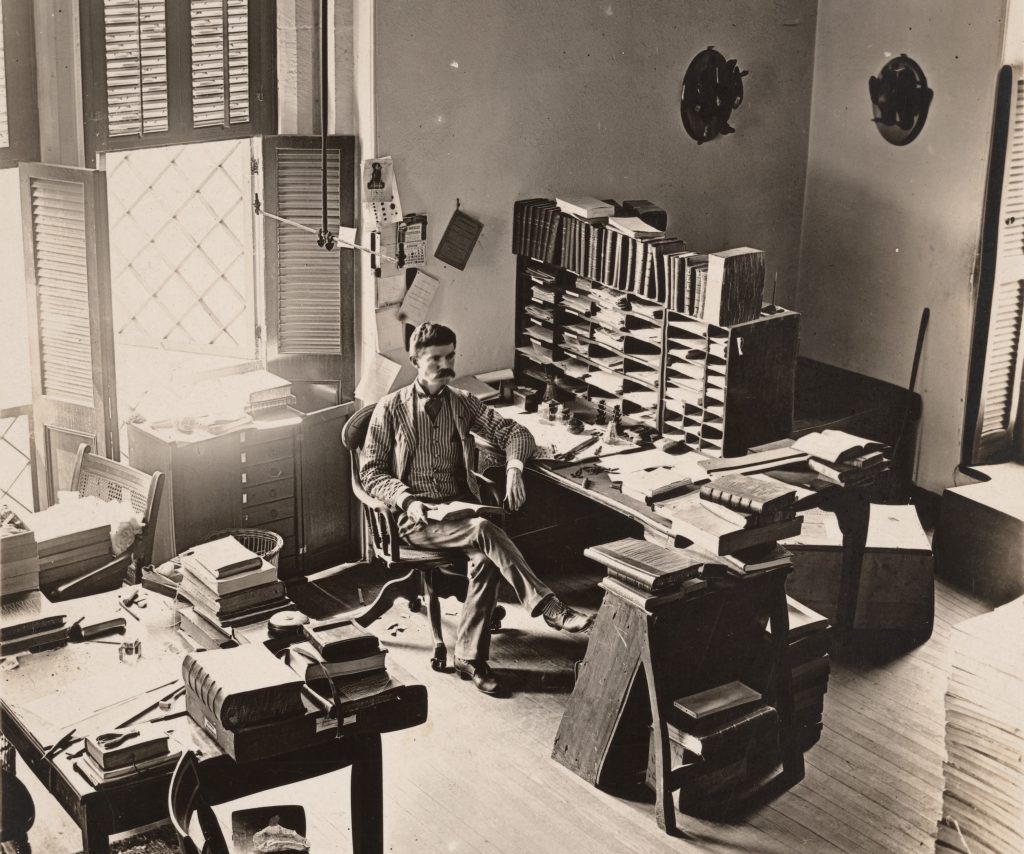 Black and white photograph of man seated in office, surrounded by desks, books and files. 