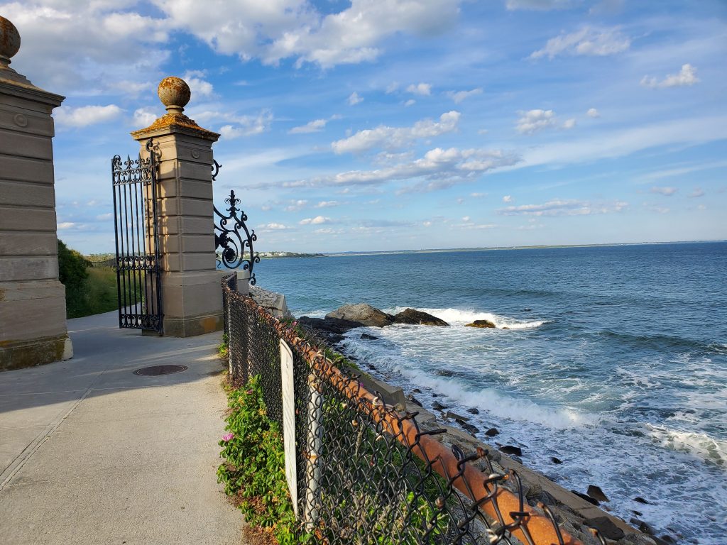 Stone pathway alongside a rocky cliff with a view of rolling waves and a marble ironed fence. 