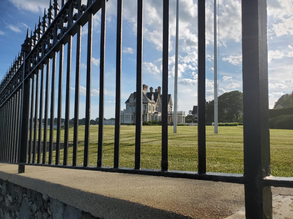 Cast iron fence gate in foreground with a cloud filled sunny sky with a gleaming white mansion The Breakers in the far distant background 