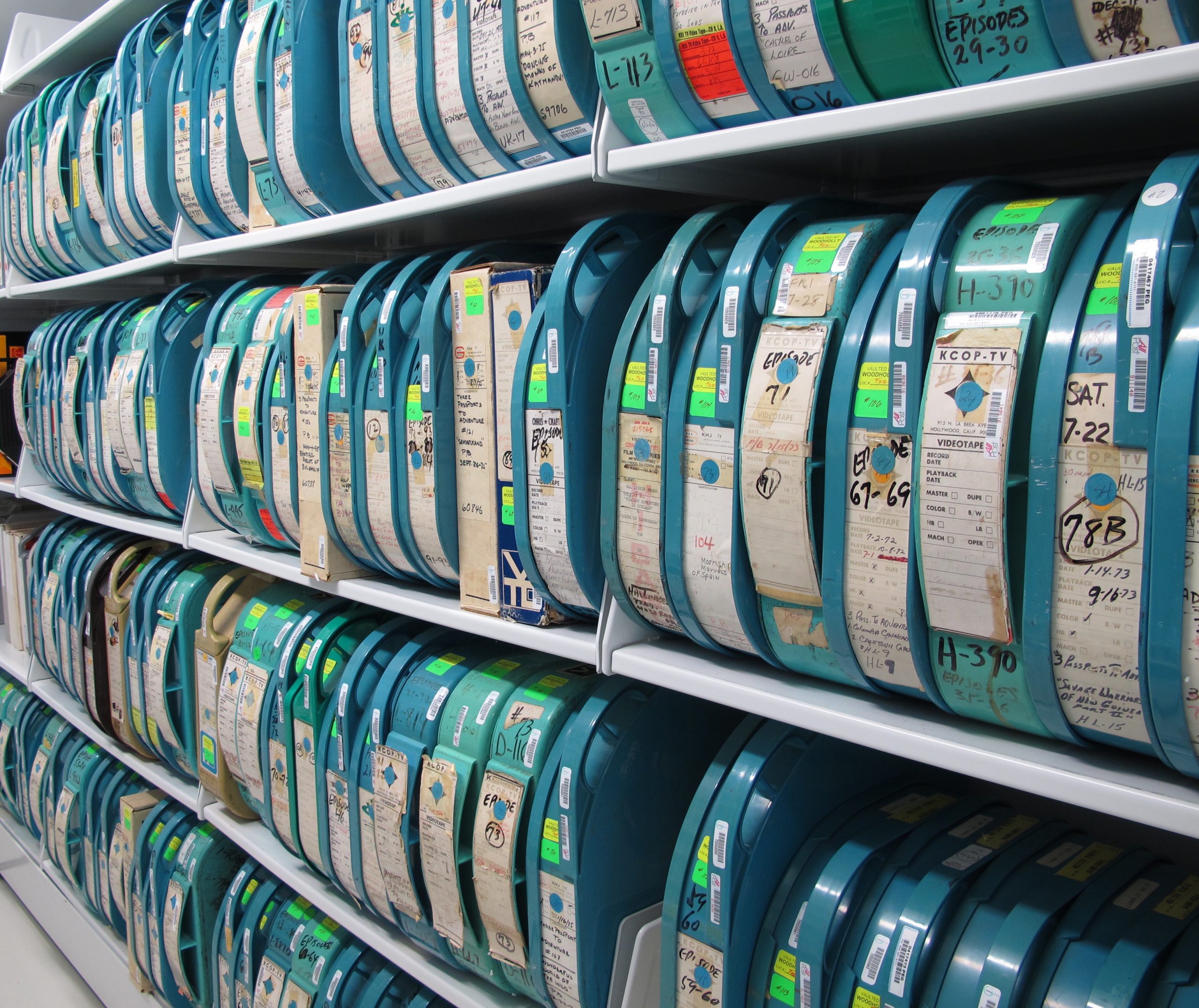 Blue plastic containers of Quadruplex videotape with varying labels and colored stickers sit on four rows of upright shelving.