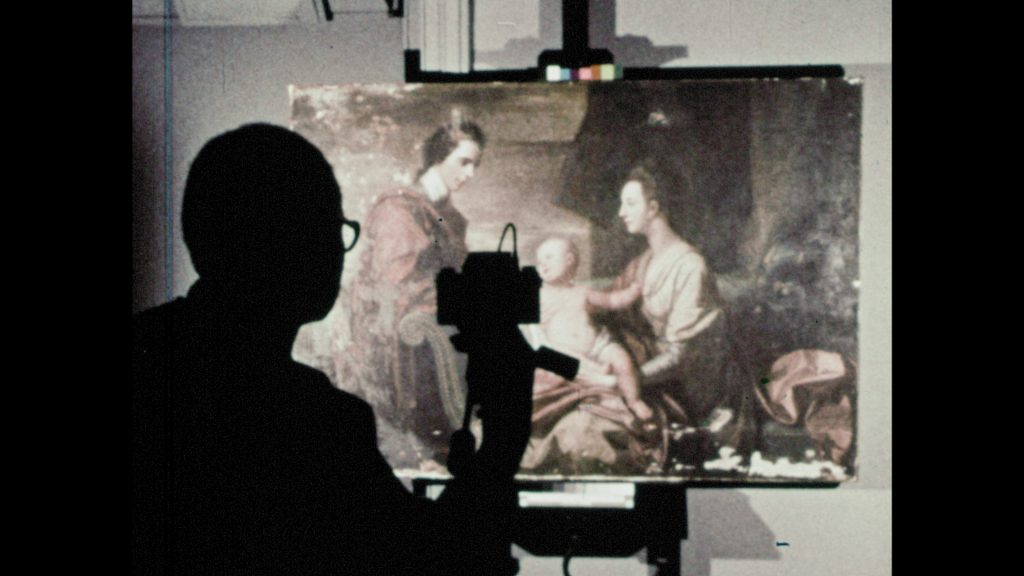 A person in the foreground sits in the shadows, while studio lights illuminate a painting of a family in the background, as a still image camera is pointed to take a photograph.