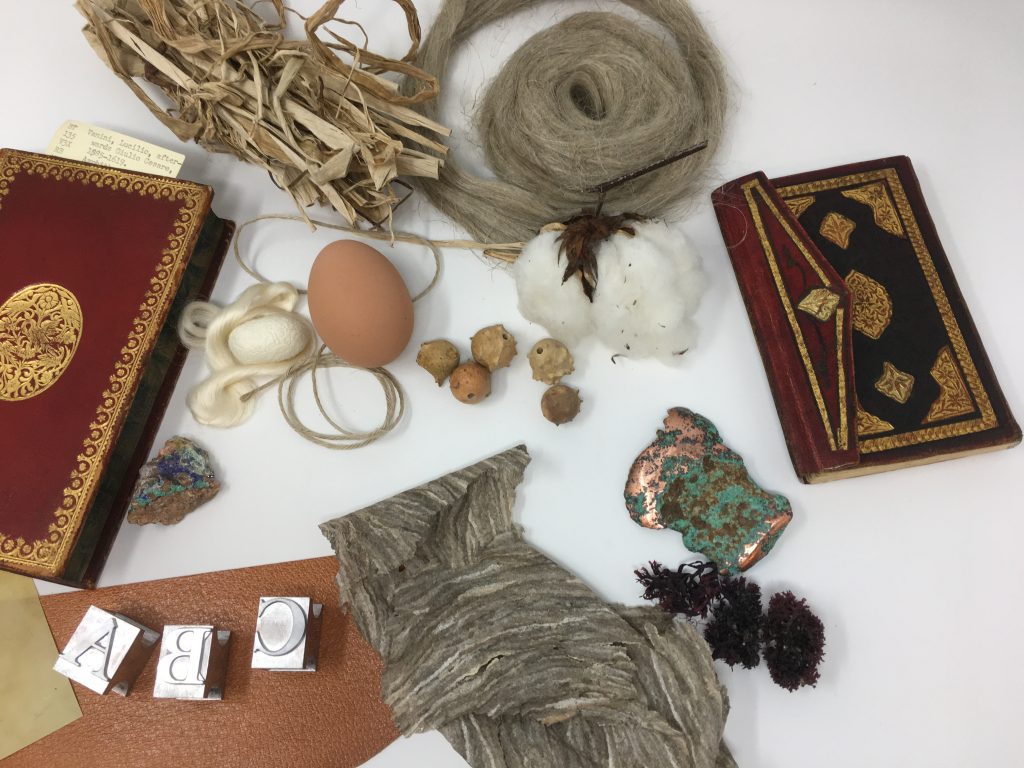 A table top is covered with materials used to make books. The include a waspnest, type, an egg, and other natural materials. A small book is on either side. 