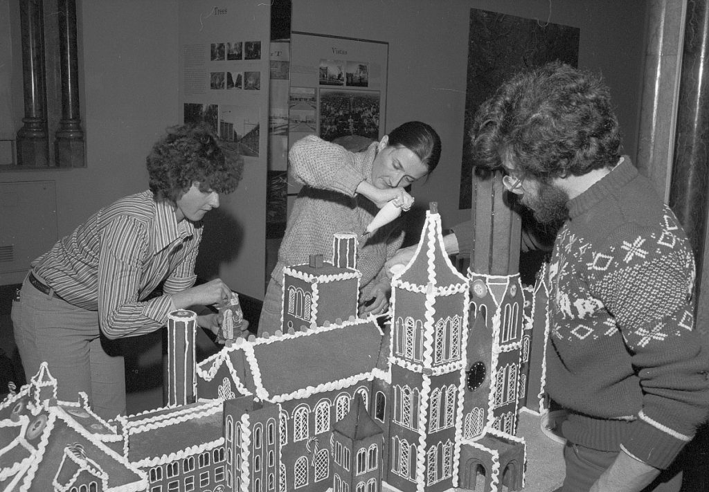 Black and white photo of three people decorating a giant gingerbread house shaped like the Smithsonian Castle.