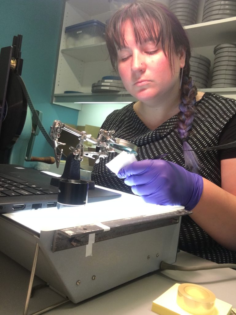 Emily Nabasny wears purple nitrile gloves and inspects a piece of film under magnifying equipment.