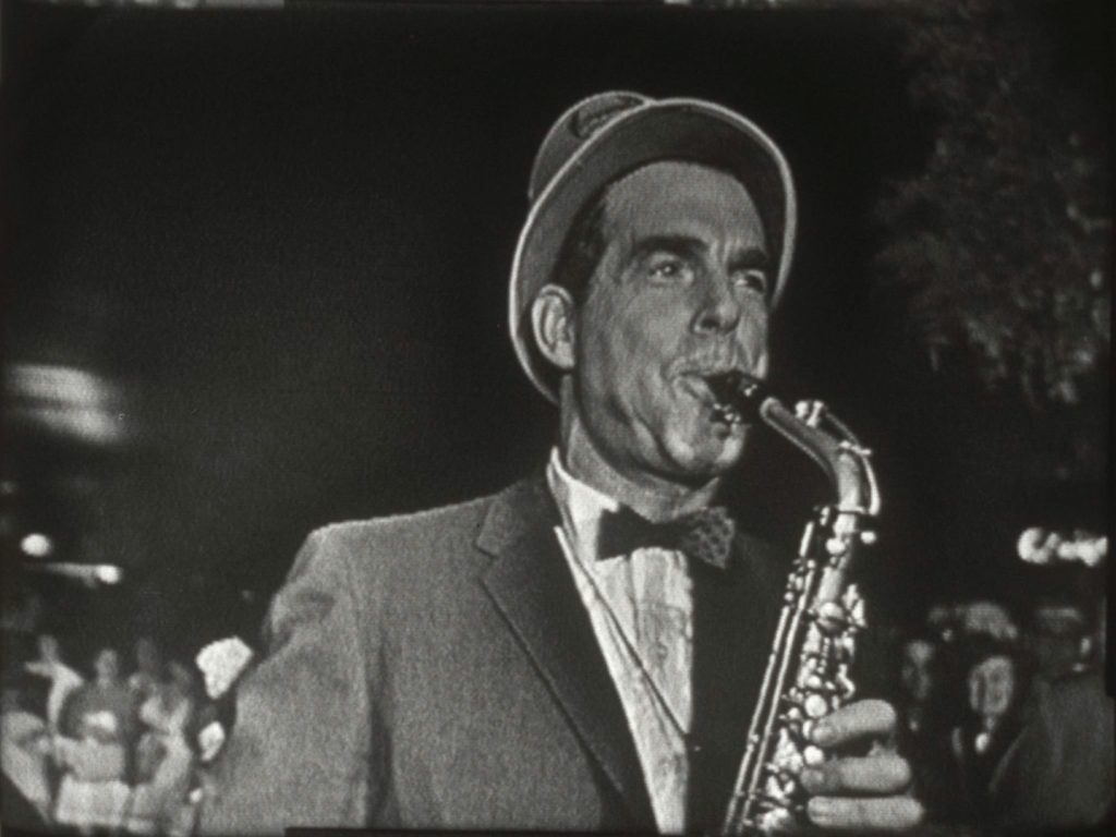 Actor Fred MacMurray wears a hat and bow tie, while blowing on a saxophone.