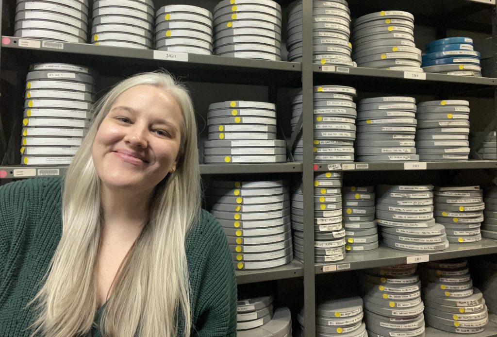 Analiese Oetting smiles in a green sweater, standing in front of a shelf of hundreds of motion picture film cans.