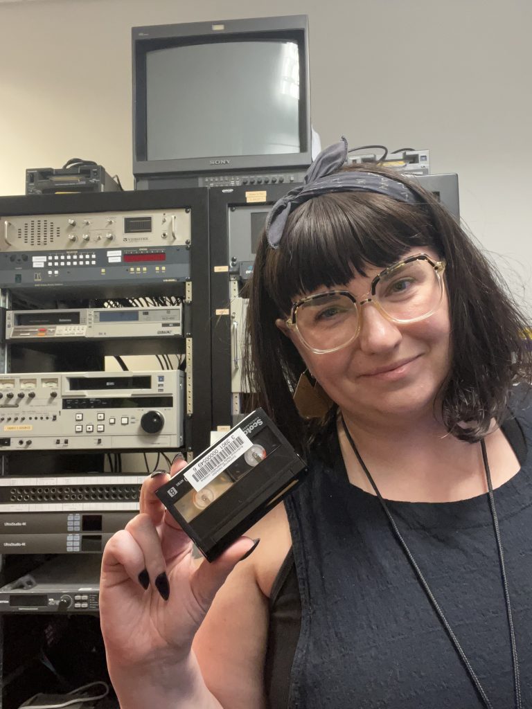 Brianna Toth holds a Video8 tape in front of a rack of av equipment.