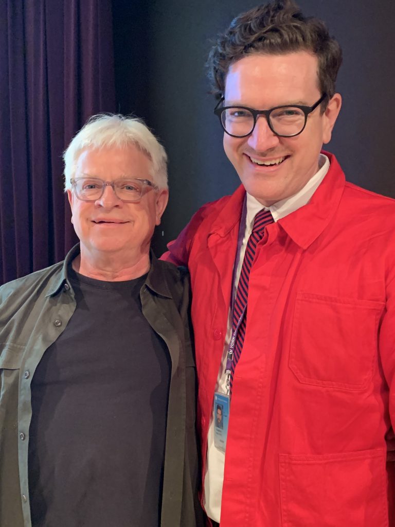 A man with white hair and black shirt smiles at left, while a man in red jacket and tie smiles at right.