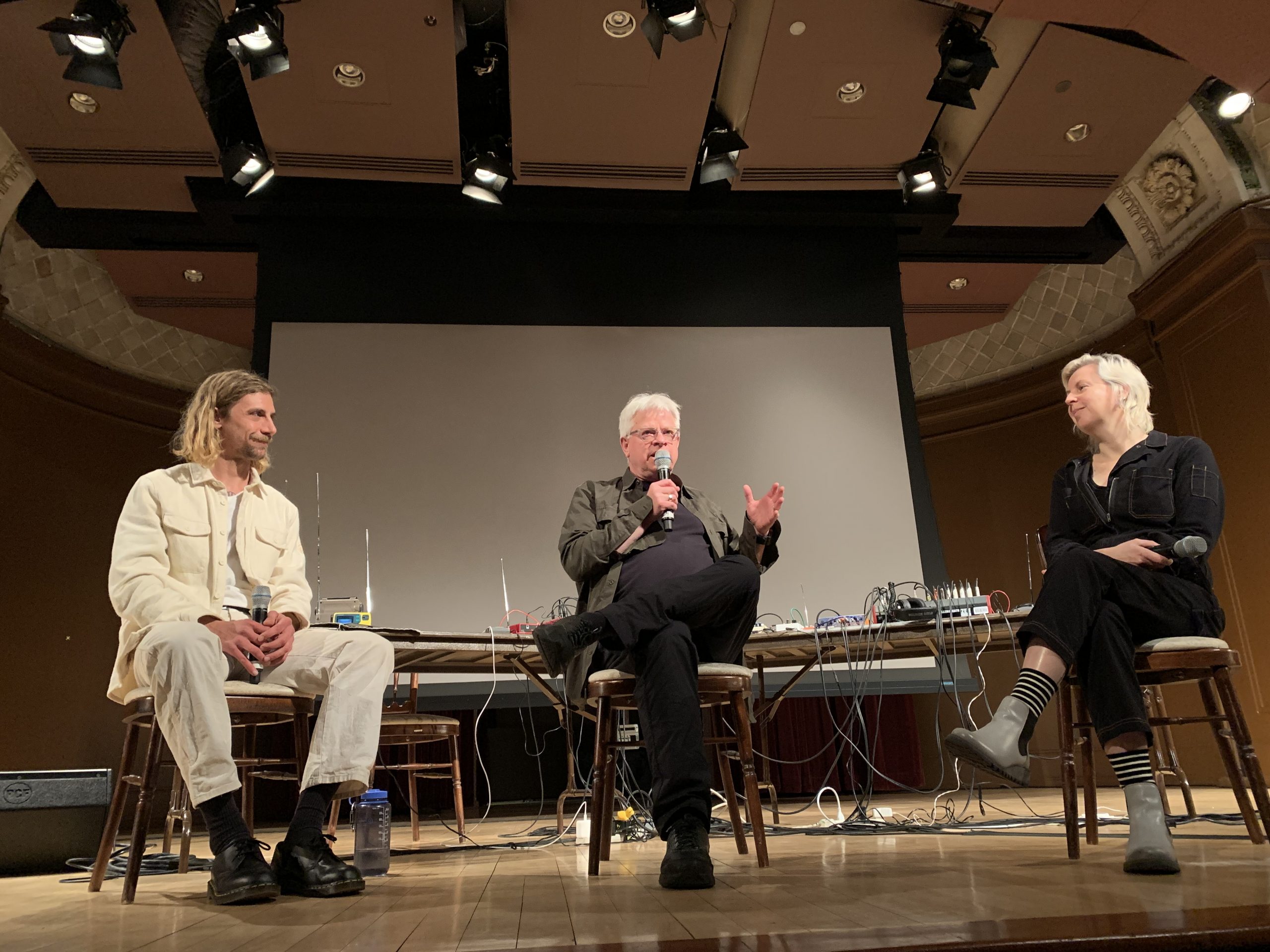 Three people sit on chairs atop the stage of the Baird Auditorium, in conversation. The middle speaker, Rick Prelinger, holds a microphone and gestures with his opposing hand.