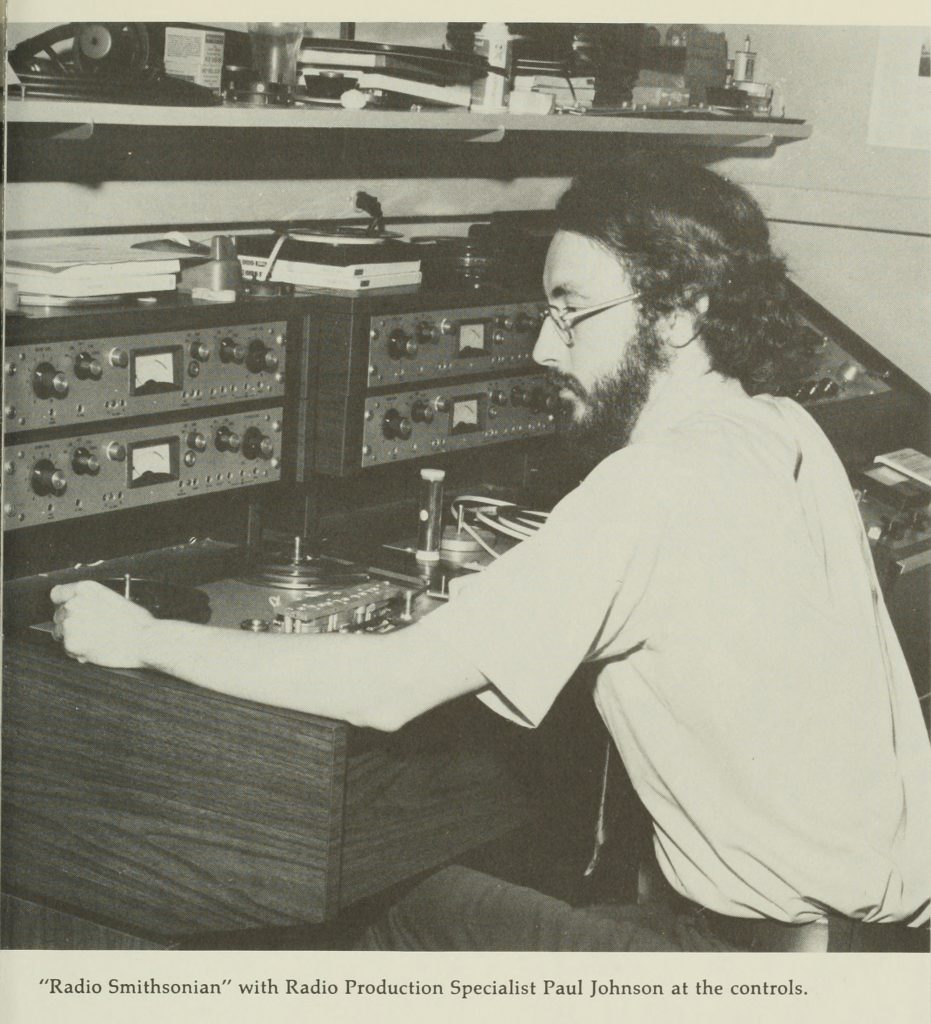 A bearded man leans over two audiotape recording decks, winding the end of a tape onto a take-up reel.