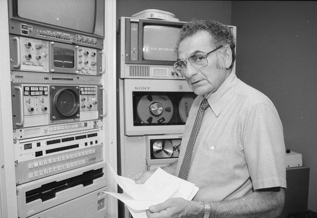 A man in glasses and short-sleeved shirt with tie holds papers, standing in front of racks of analog video equipment and looking directly at the camera.